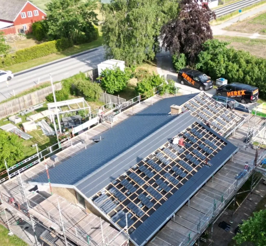 Pågående takläggning på villa med byggställning och Herex servicebilar, drönarvy